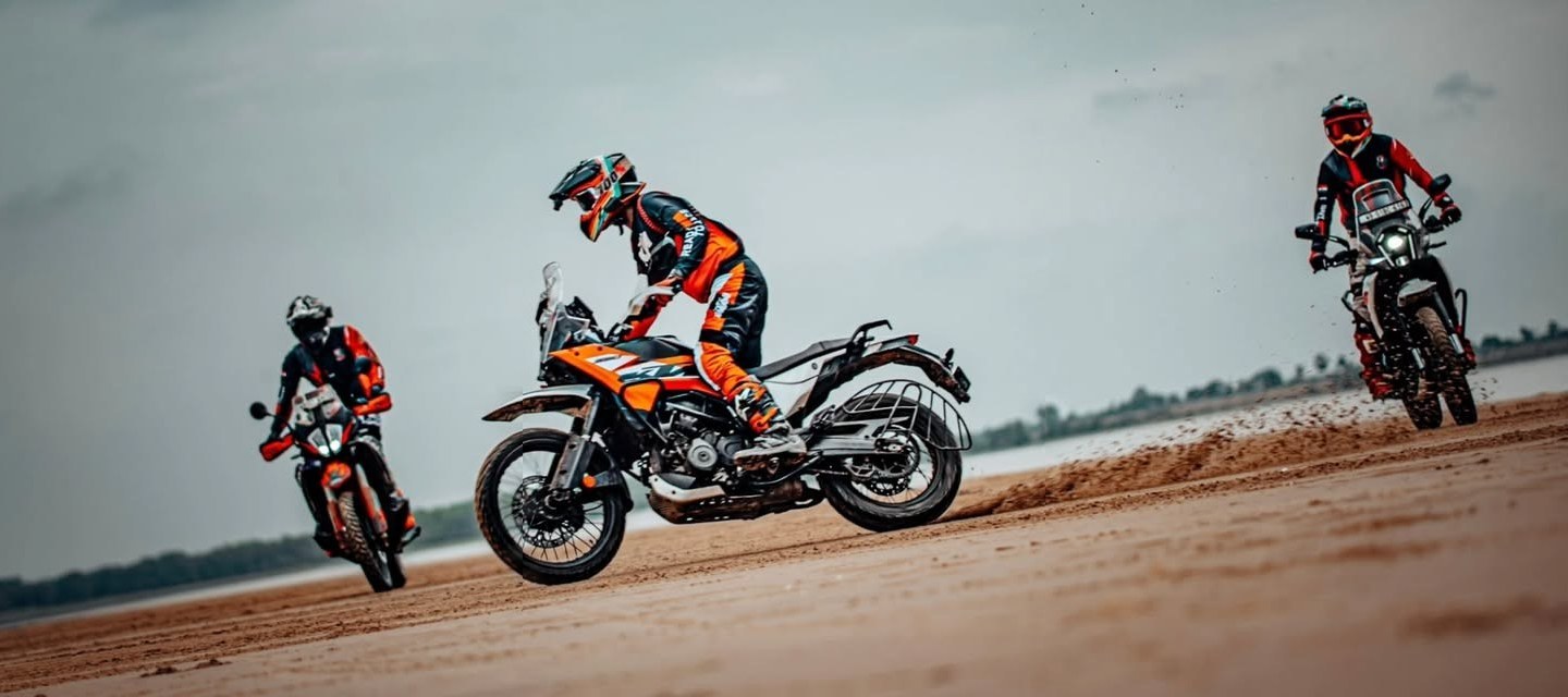 Three adventure riders carving across a sand flat, roost flying
